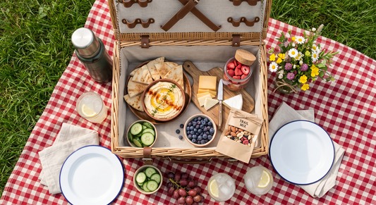 A pantry-powered picnic basket on a red-and-white checkered blanket with a bowl of hummus, warm pita, freeze-dried strawberries and blueberries, hard cheese, trail mix, cucumber, grapes, and lemonade