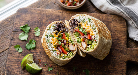 Overhead view of a sliced burrito filled with rice, beans, chicken, and vegetables on a wooden cutting board with lime wedges and cilantro